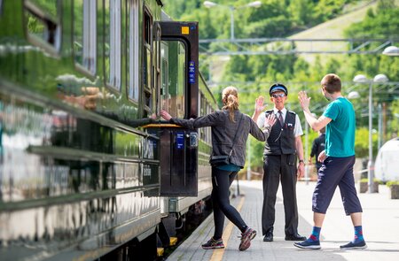 Zwei Personen steigen nacheinander in einen Zug ein und winken dem Schaffner zum Abschied zu.  | © Sverre Hjornevik
