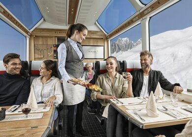 Zwei Paare sitzten im Speisewagen 2. Klasse mit Panoramafenstern und eine Mitarbeiterin bringt essen | © Glacier Express AG/Stefan Schlimpf