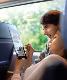 eine Person, die in einem Zug sitzt und auf ein Tablet schaut, auf dem Text und Bilder zu sehen sind.  | ©  Deutsche Bahn AG
