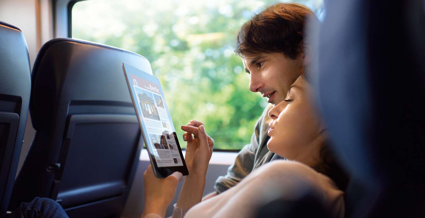  eine Person, die in einem Zug sitzt und auf ein Tablet schaut, auf dem Text und Bilder zu sehen sind.  | ©  Deutsche Bahn AG