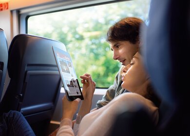  eine Person, die in einem Zug sitzt und auf ein Tablet schaut, auf dem Text und Bilder zu sehen sind.  | ©  Deutsche Bahn AG
