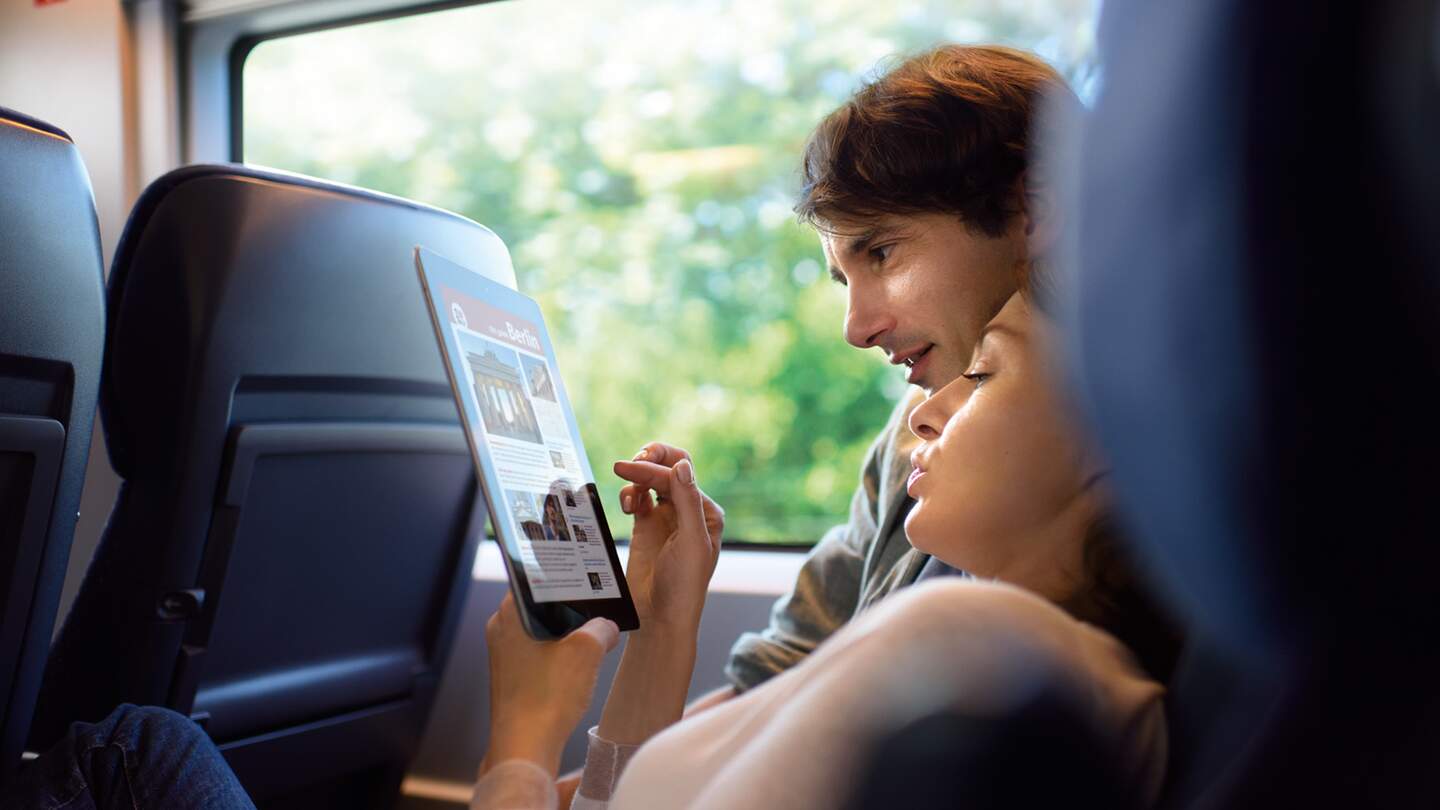  eine Person, die in einem Zug sitzt und auf ein Tablet schaut, auf dem Text und Bilder zu sehen sind.  | ©  Deutsche Bahn AG