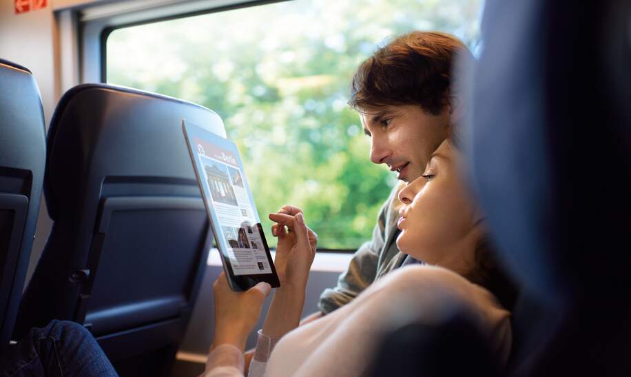  eine Person, die in einem Zug sitzt und auf ein Tablet schaut, auf dem Text und Bilder zu sehen sind.  | ©  Deutsche Bahn AG