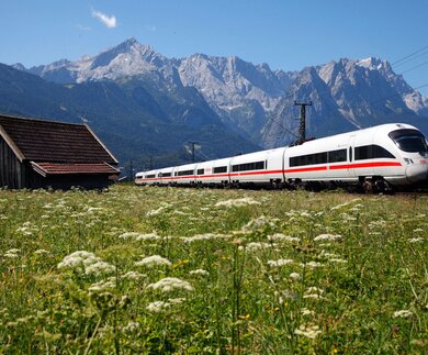 Ein moderner weißer Zug mit roten Akzenten fährt auf den Gleisen an einer Holzhütte vorbei, vor der Kulisse majestätischer Berge unter einem klaren blauen Himmel. | © Deutsche Bahn AG