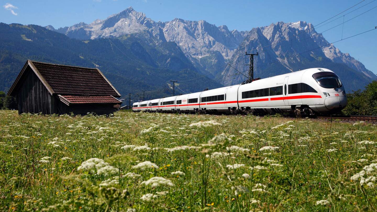 Ein moderner weißer Zug mit roten Akzenten fährt auf den Gleisen an einer Holzhütte vorbei, vor der Kulisse majestätischer Berge unter einem klaren blauen Himmel. | © Deutsche Bahn AG