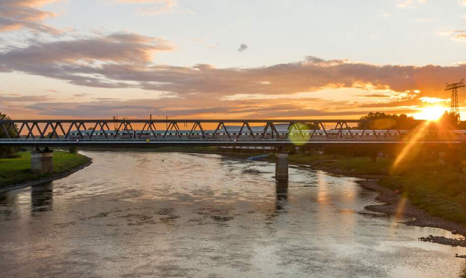 Eine Eisenbahnbrücke überspannt einen Fluss bei Sonnenuntergang, wobei das warme Licht der Sonne auf der Wasseroberfläche reflektiert. | ©  Deutsche Bahn AG