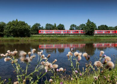 einen rot-weißen Zug, der entlang von Gleisen faehrt, die parallel zu einem Gewaesser verlaufen, wobei sich der Zug im Wasser spiegelt. | © Deutsche Bahn AG/Bartlomiej Banaszak