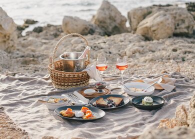 ein Picknick-Setup an einem Sandstrand mit Felsen im Hintergrund,darunter  ein Weidenkorb, Teller mit Essen, Besteck, Weinglaese | © gettyimages.com/Polina Lebed