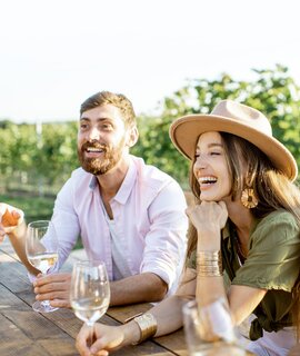 Menschen, die Wein trinken und sich miteinander unterhalten, waehrend sie an einem sonnigen Abend am Esstisch im Freien auf dem Weinberg sitzen | © Gettyimages.com/RossHelen