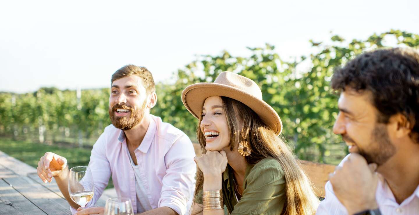 Menschen, die Wein trinken und sich miteinander unterhalten, waehrend sie an einem sonnigen Abend am Esstisch im Freien auf dem Weinberg sitzen | © Gettyimages.com/RossHelen