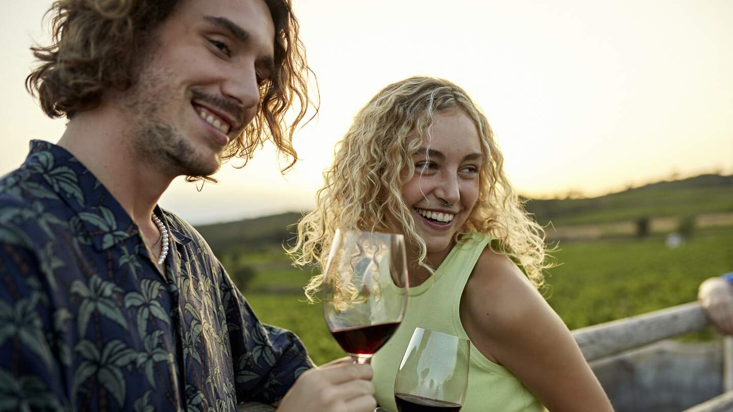 Nahaufnahme von Urlaubern in Freizeitkleidung, die Rotweinglaeser halten und am spaeten Nachmittag von der Kamera mit landwirtschaftlichem Feld im Hintergrund wegschauen. | © Gettyimages.com/xavierarnau