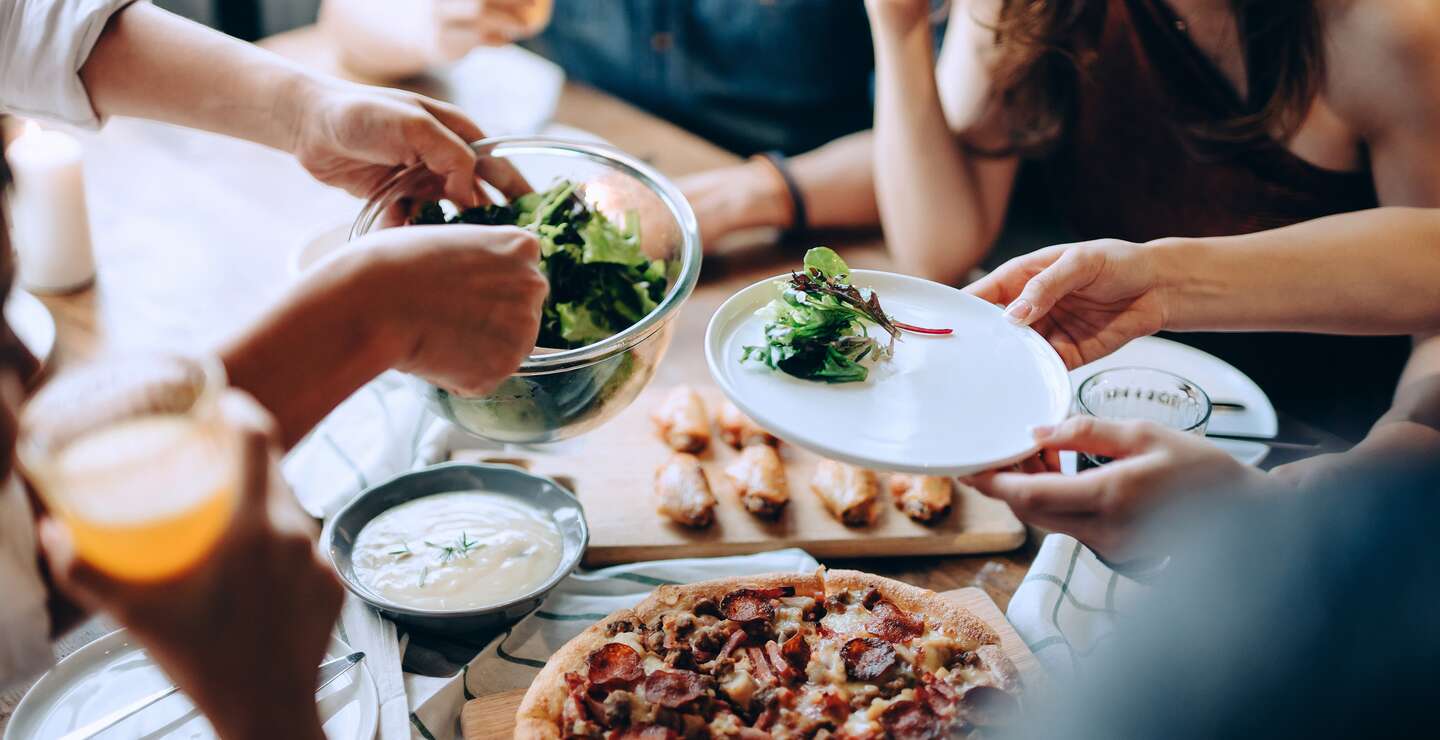Nahaufnahme einer jungen Gruppe froehlicher Freunde, die Spass daran haben, zusammen zu genießen, Essen ueber den Esstisch auf der Party zu verteilen und zu teilen | © Gettyimages.com/AsiaVision