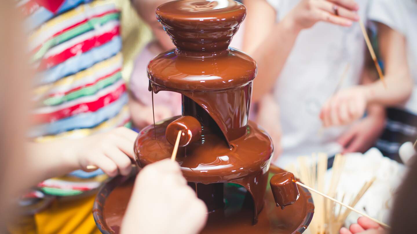 Lebendiges Bild von Chocolate Fountain Fontain auf einer Kindergeburtstagsparty mit herumspielenden Kindern und Marshmallows und Fruechten, die in den Brunnen tauchen | © Gettyimages.com/tsguliev
