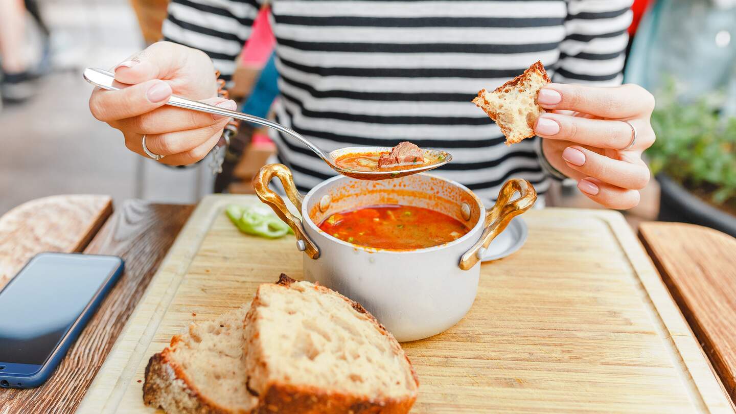 Eine Frau isst eine traditionelle ungarische Gulaschsuppe aus einem Topf in einem Restaurant im Freien | © Gettyimages.com/frantic00