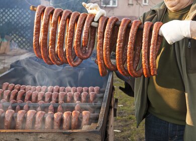 Mann haelt eine Stange mit traditionellen Raeucherwuersten in die Hoehe, dahinter befinden sich weitere Raeucherwuerste | © Gettyimages.com/Avalon_Studio