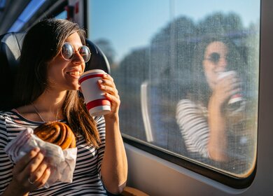 Person sitzt im Zug und hält einen Kaffeebecher in der einen Hand und ein Gebäck in der anderen, mit Blick aus dem Fenster auf die vorbeiziehende Landschaft. | © GettyImages.com/	urbazon