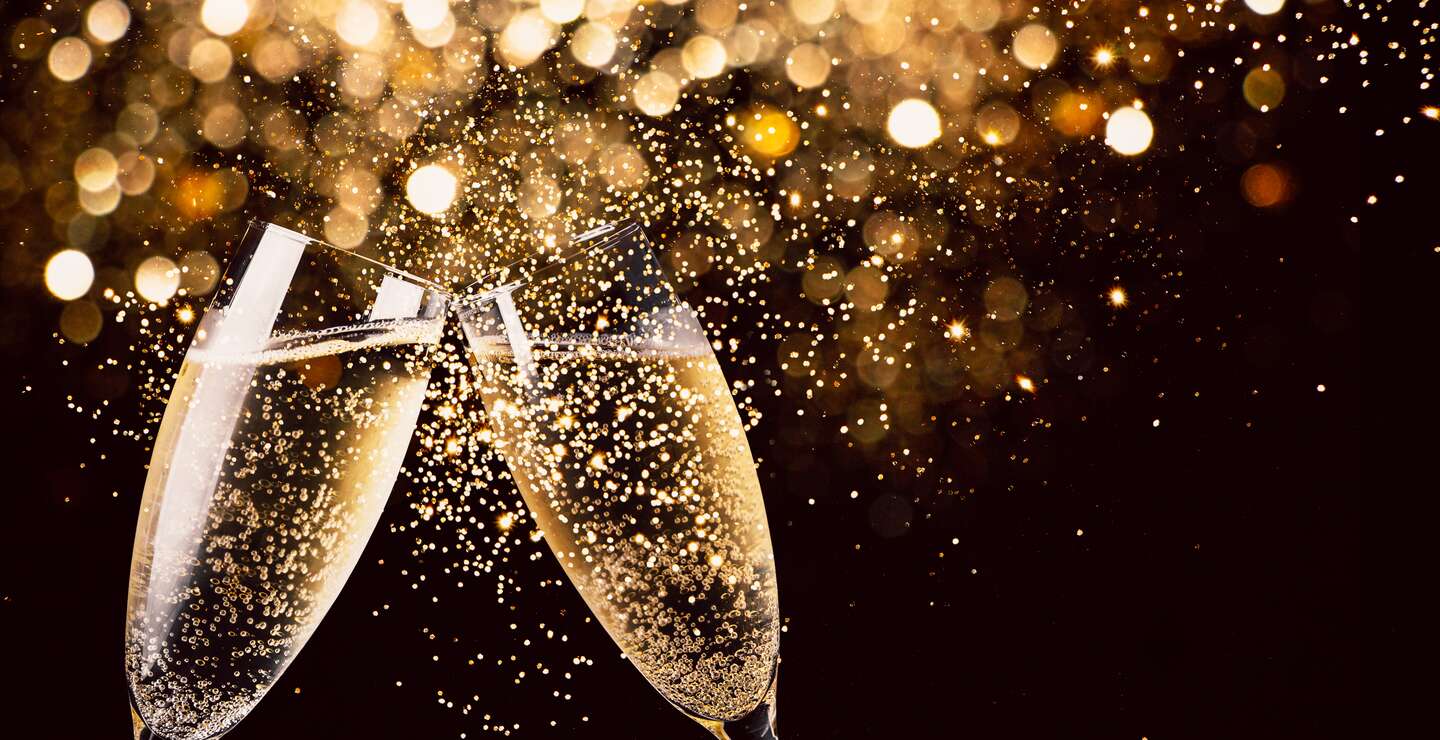 zwei Glaeser Champagner stossen in der Nacht mit Lichtern im  Glitzer und Funken im Hintergrund an | © Gettyimages.com/ASIFE