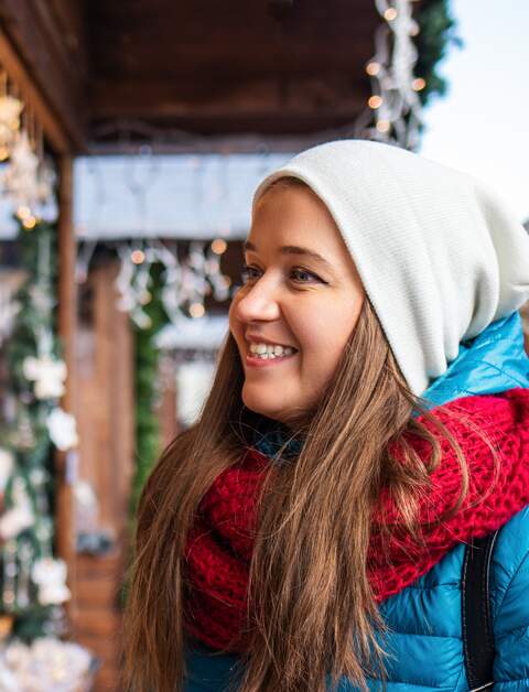 Froehliche junge Frau betrachtet bunte Weihnachtsschmuck-Auslagen an einem Holzhuettestand auf dem Weihnachtsmarkt bei schoenem Winterwetter | © Gettyimages.com/Zbynek Pospisil
