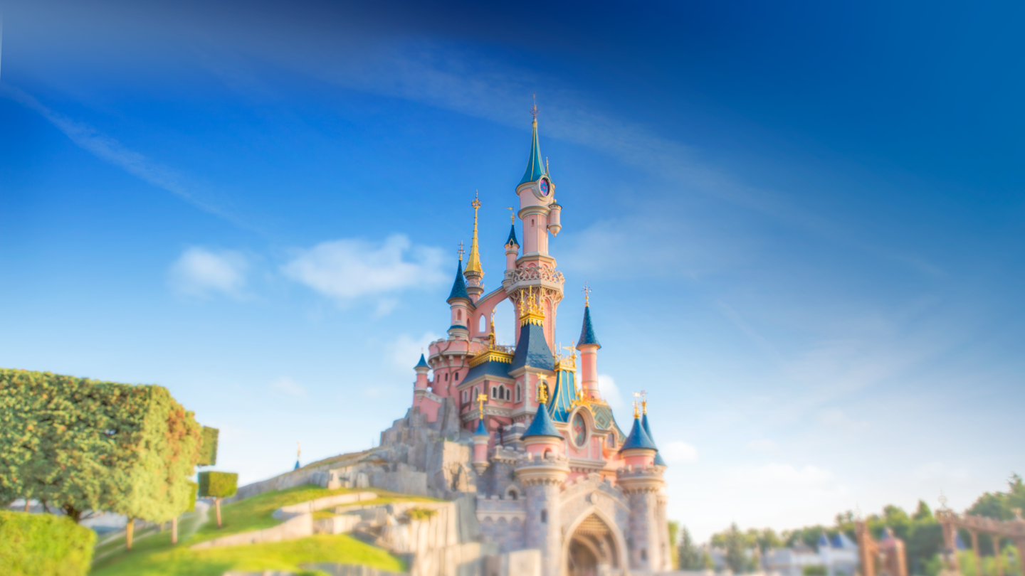 Das rosane Sleeping Beauty Castle mit blauen Tuermen, Fotografiert vom Central Plaza, umgeben von bunten Blumen. | © Disney