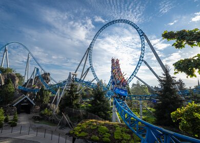 Blaue Loopingachterbahn rast bei sonnigem Wetter durch hohe Kurven und über grüne Landschaft, während die Fahrgäste den intensiven Adrenalinkick erleben. | © Europa-Park