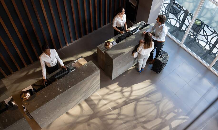 Junges Geschaeftspaar, das im Hotel eincheckt und schoene Rezeptionistin, die ihnen sehr froehlich hilft | © Gettyimages.com/Hispanolistic