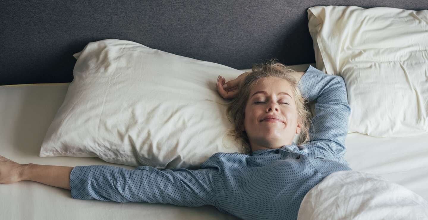 Laechelnde junge Frau, die an einem sonnigen Morgen ihre Arme im Bett streckt | © Gettyimages.com/FreshSplash