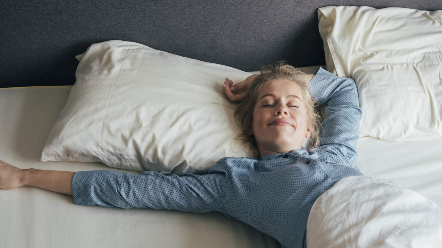 Laechelnde junge Frau, die an einem sonnigen Morgen ihre Arme im Bett streckt | © Gettyimages.com/FreshSplash