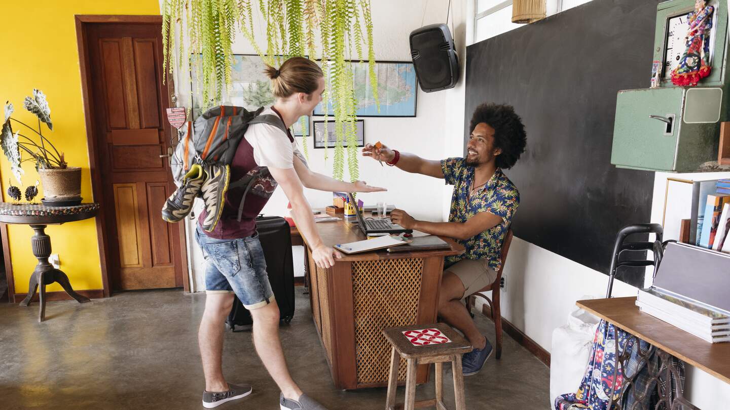 Junger Rucksacktourist checkt im Hostel ein, Gap-Year-Student nimmt Mann, der an der Rezeption arbeitet, Schlüssel ab | © Gettyimages.com/JohnnyGreig