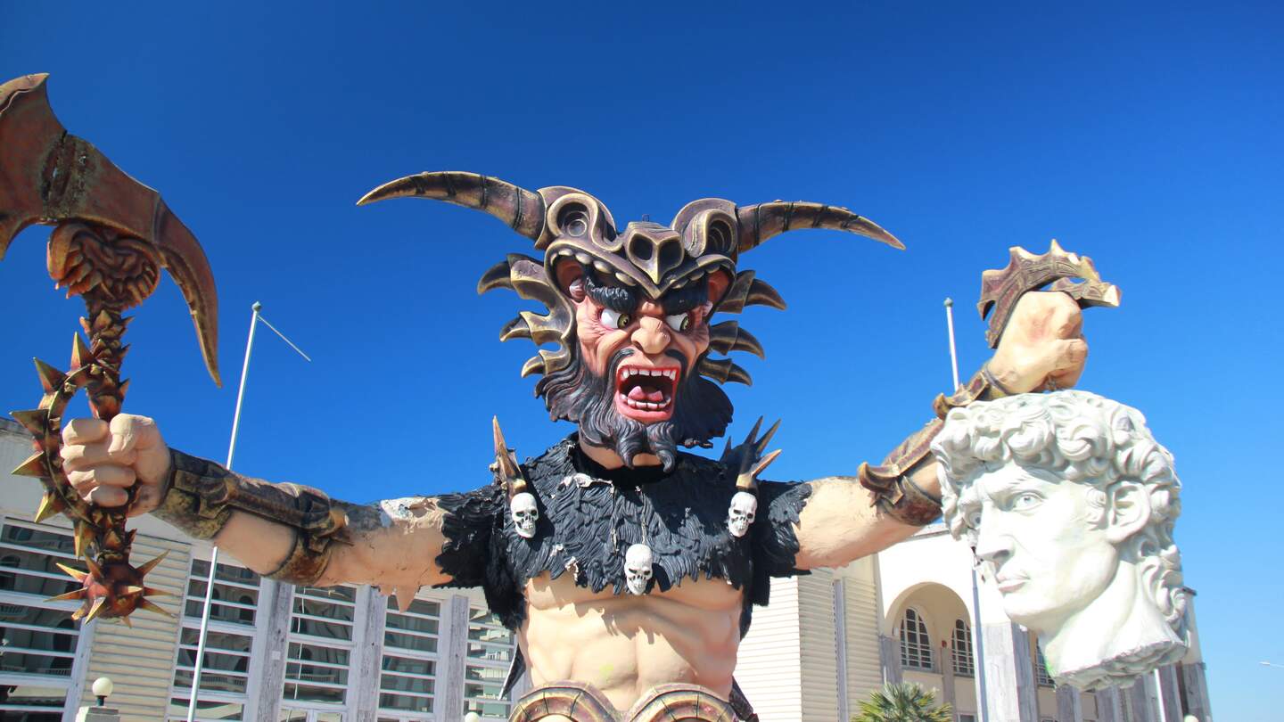 Eine große Pappmaché-Figur in Form eines gehörnten Dämons mit Beil und abgeschlagenen Kopf in jeweils einer Hand vor blauem Himmel beim Faschingsumzug in Viareggio, Italien | © aleperini/pixabay