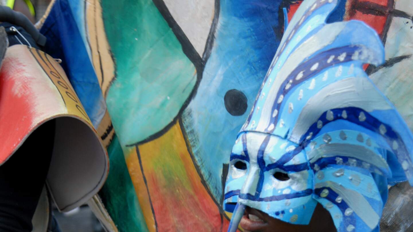 Eine farbige Frau hält sich beim Umzug eine blaue sebstgestaltete Halbmaske aus Pappe vor ihr Gesciht beim Nottingg Hill Karneval in London | © GettyImages.com/grahamheywood