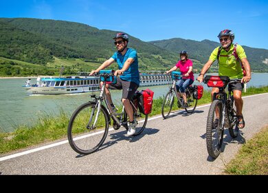 Drei Radfahrer im Sommer neben dem Fluss, im Hintergrund ein Flusskreuzfahrtschiff | © Carina Eßbüchl