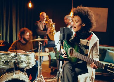 Eine Frau singt und spielt Gitarre, während sie auf einem Stuhl mit verkreuzten Beinen sitzt. Im Hintergrund sind weitere Bandmitglieder. | © Gettyimages.com/millann
