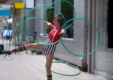 An einem heissen Tag in der Stadt dreht eine Strassenkuenstlerin Reifen um ihre Arme und Beine, waehrend sie auf einem Bein balanciert. Sie traegt ein gestreiftes, farbenfrohes Kostuem | © Gettyimages.com/Vladimir Vladimirov