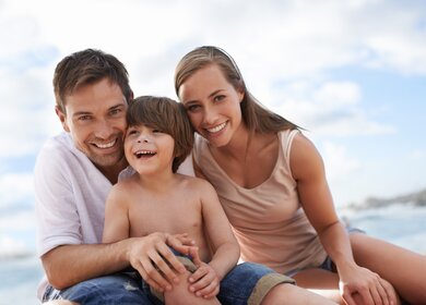 Die Eltern sitzen mit ihrem Sohn am Strand und umarmen sich | © Gettyimages.com/PeopleImages