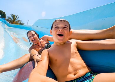 Sohn und Mutter, die lachen und schreien, waehrend sie schnell auf einer Wasserrutsche im Wasserpark herunterrutschen | © Gettyimages.com/Basilico Studio Stock