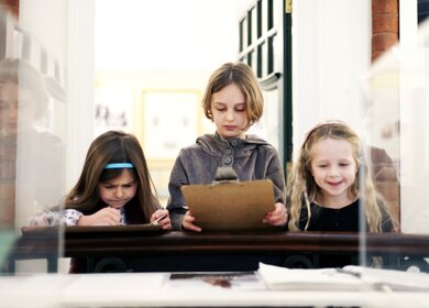 Drei Kinder in einer Museums Ausstellung stehen vor einem Glaskasten. | © Gettyimages.com/Rene Mansi