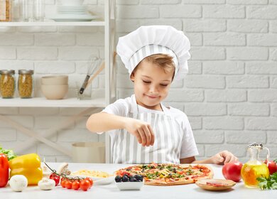 Kleiner Junge mit Kochmuetze und Schuerze kocht Pizza in der Kueche. Kind fuegt Kaese in Pizza hinzu | © Gettyimages.com/Erstudiostok