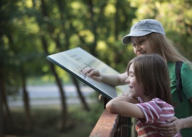 Mutter mit Kind in einer Ausstellung eines Parks oder eines Museums: Gemeinsam lesen sie eine Erklärtafel und entdecken spannende Informationen | © Gettyimages.com/portishead1