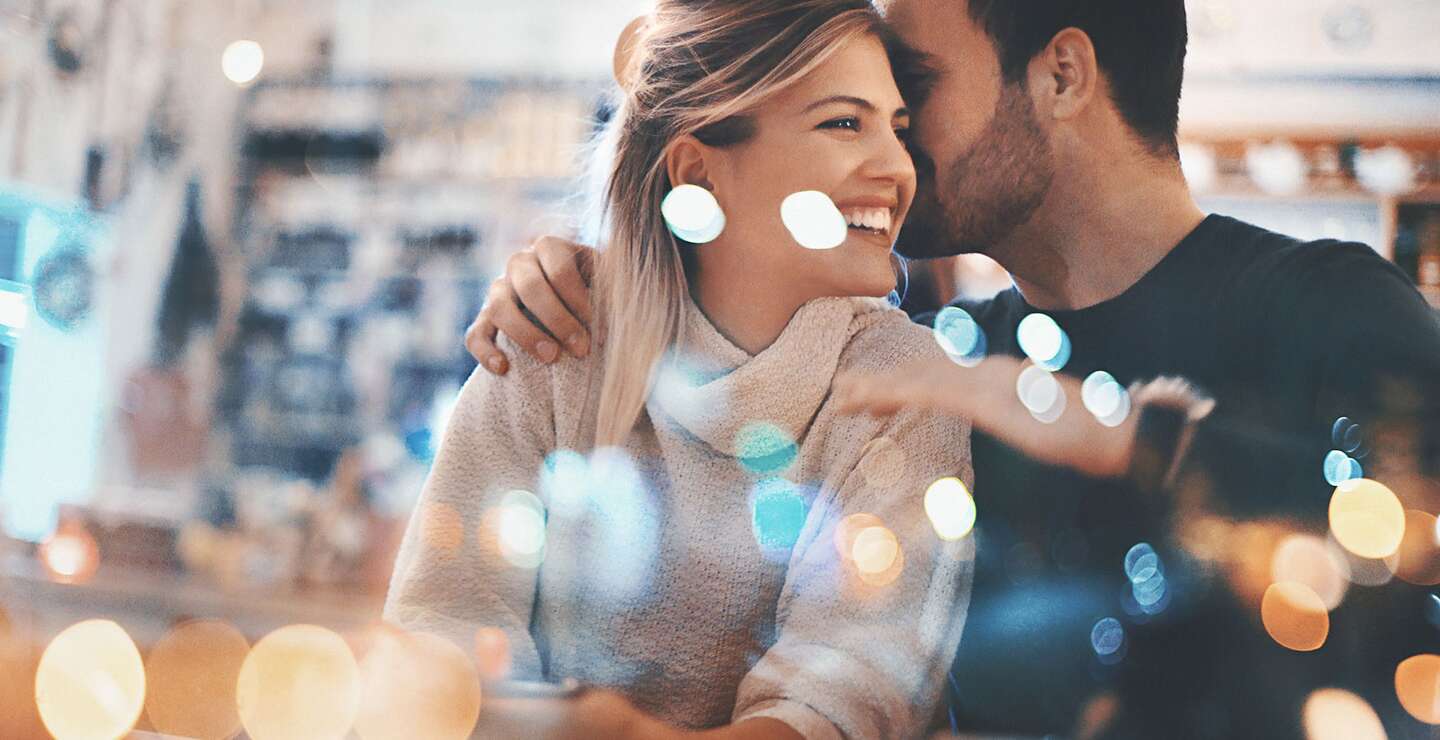 Nahaufnahme eines Paares Mitte 20, das einen romantischen Abend in einem gemütlichen Café verbringt und Spaß hat. Er flüstert ihr etwas Süßes ins Ohr und das bringt sie zum Lächeln. Von außen durch das große Fenster fotografiert, neben dem sie sitzen. Viele defokussierte, schimmernde Lichter im Rahmen. | © Gettyimages.com/gilaxia