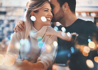 Nahaufnahme eines Paares Mitte 20, das einen romantischen Abend in einem gemütlichen Café verbringt und Spaß hat. Er flüstert ihr etwas Süßes ins Ohr und das bringt sie zum Lächeln. Von außen durch das große Fenster fotografiert, neben dem sie sitzen. Viele defokussierte, schimmernde Lichter im Rahmen. | © Gettyimages.com/gilaxia