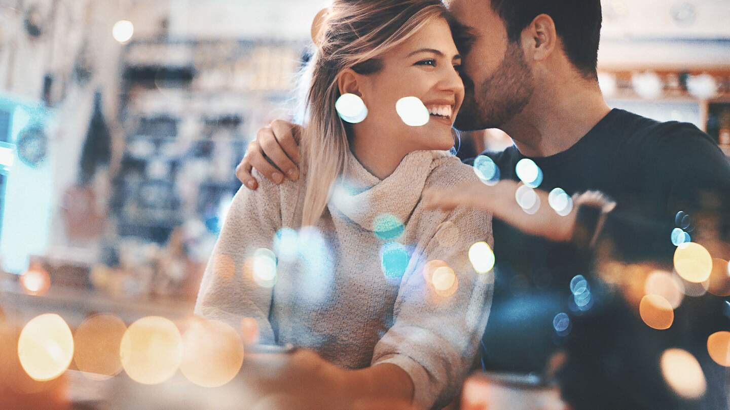 Nahaufnahme eines Paares Mitte 20, das einen romantischen Abend in einem gemütlichen Café verbringt und Spaß hat. Er flüstert ihr etwas Süßes ins Ohr und das bringt sie zum Lächeln. Von außen durch das große Fenster fotografiert, neben dem sie sitzen. Viele defokussierte, schimmernde Lichter im Rahmen. | © Gettyimages.com/gilaxia