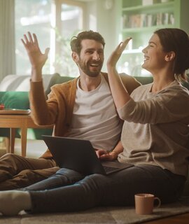 Paar benutzt Laptopund gibt sich ein High-Five, während sie im Wohnzimmer auf dem Fussboden sitzen | ©  Gettyimages.com/gorodenkoff