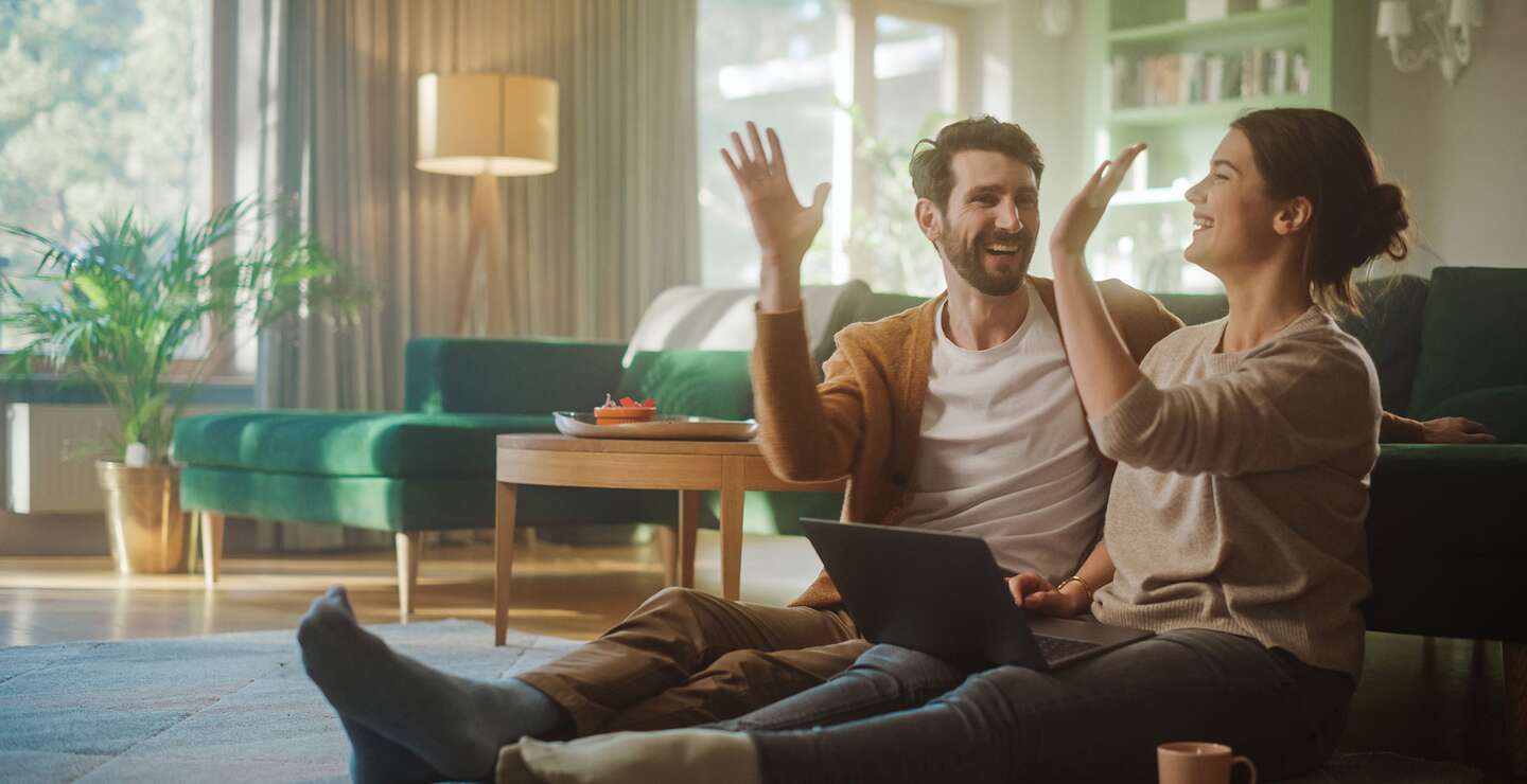 Paar benutzt Laptopund gibt sich ein High-Five, während sie im Wohnzimmer auf dem Fussboden sitzen | ©  Gettyimages.com/gorodenkoff