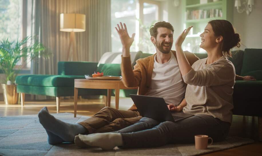 Paar benutzt Laptopund gibt sich ein High-Five, während sie im Wohnzimmer auf dem Fussboden sitzen | ©  Gettyimages.com/gorodenkoff