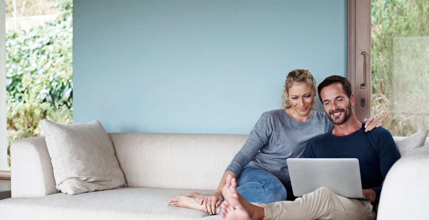 Ganzkoerperaufnahme eines Paares, das einen Laptop benutzt, während es sich auf dem Sofa entspannt | © kupicoo/GettyImages