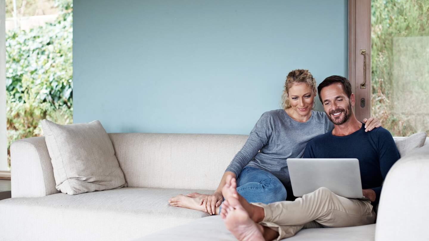 Ganzkoerperaufnahme eines Paares, das einen Laptop benutzt, während es sich auf dem Sofa entspannt | © kupicoo/GettyImages