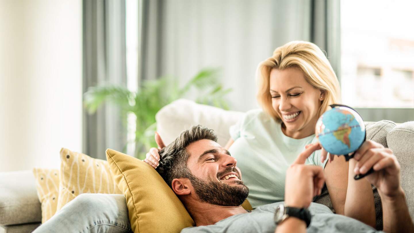 Glueckliches junges Paar plant ihre neue Reise. Junger Mann entspannt sich mit seiner Freundin auf dem Sofa und blickt auf den Globus. | © GettyImages.com/DjelicS