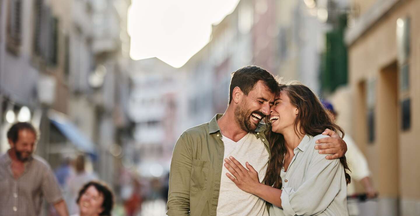 Ein Paar geht durch eine sonnige Altstadtstraße mit historischen Gebäuden, engen Gassen und Laternen, während im Hintergrund Menschen flanieren. | © Gettyimages.com/skynesher