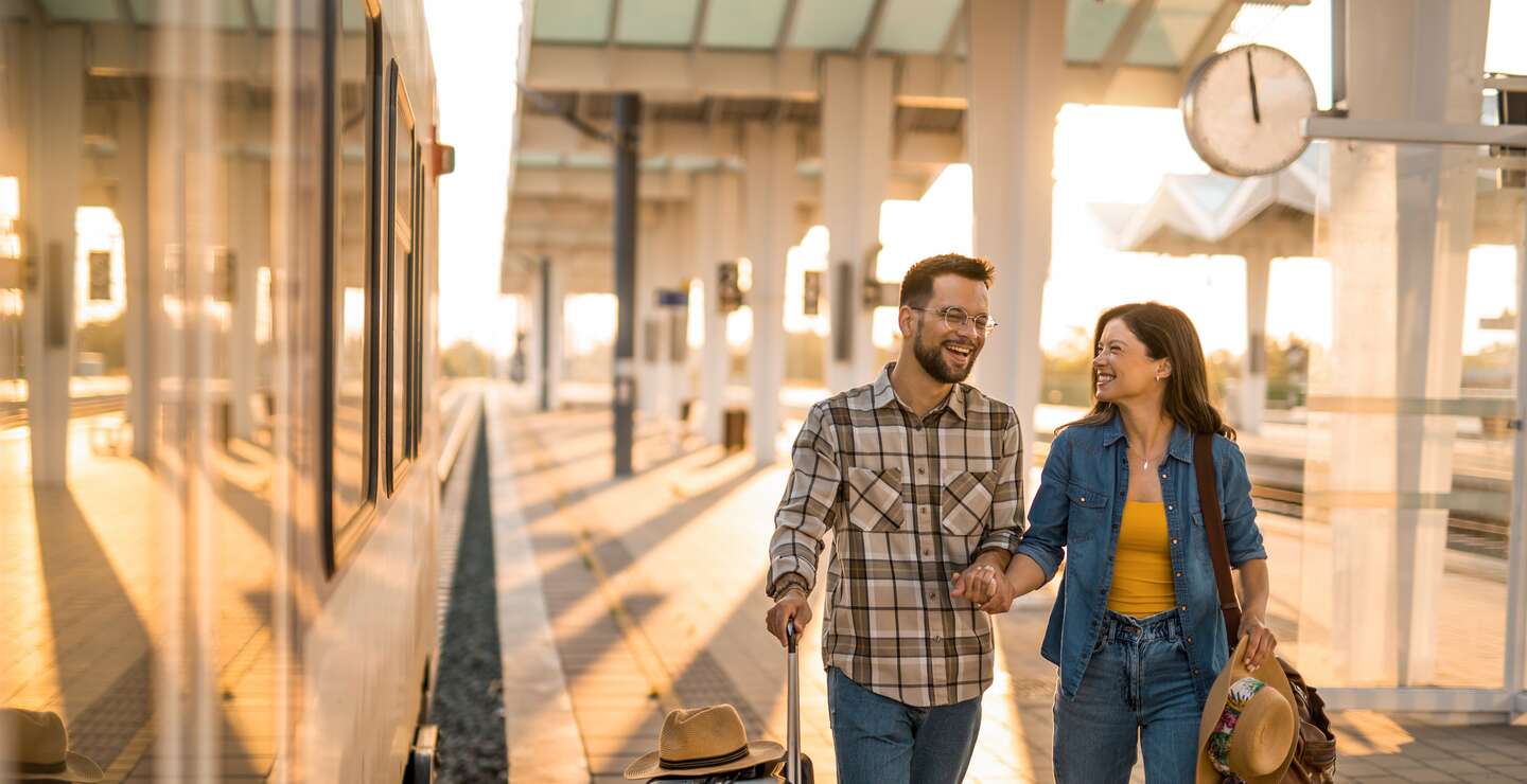 Mann und Frau laufen lachend einen Bahnsteig mit Koffer und Reisetasche entlang | © Gettyimages.com/DjelicS