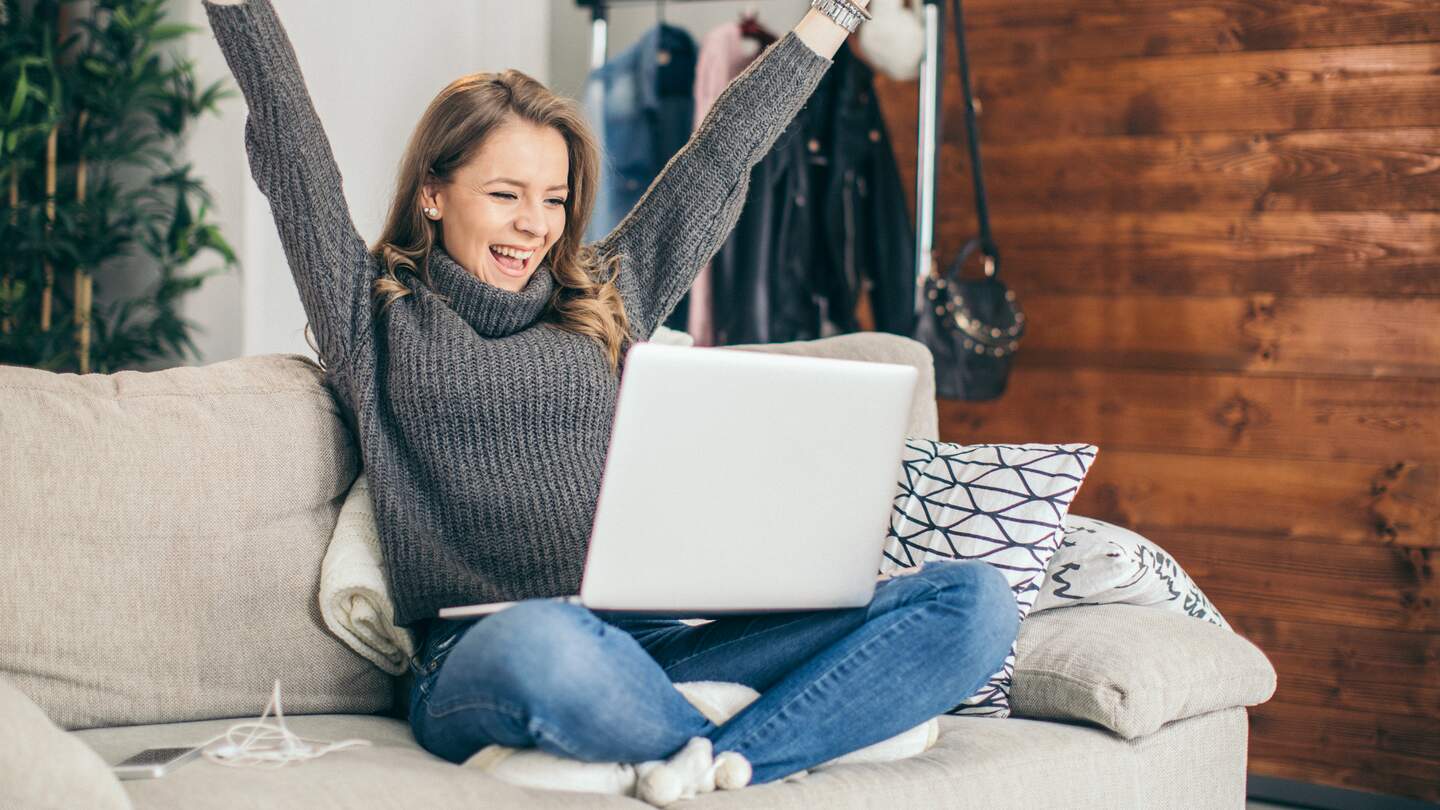 Frau sitzt auf einem Sofa mit erhobenen Armen und schaut freudig auf einen Laptop auf dem Schoß; im Hintergrund eine Holzvertäfelung und hängende Kleidung. | © Gettyimages.com/supersizer